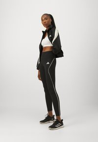 Giacca sportiva nera con accenti bianchi, reggiseno sportivo bianco, leggings neri a vita alta con piping bianco e sneakers nere con tre strisce.