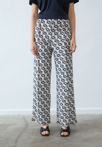 Pantalons amples et légers avec un motif géométrique noir et blanc en zigzag. Présentent une taille haute et des jambes larges, portés avec des sandales à talons noires.