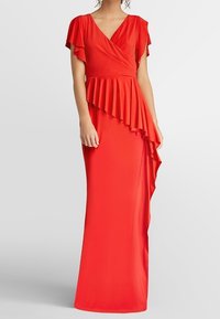 Femme portant une robe longue rouge vif avec des manches flottantes et un détail volé asymétrique à la taille et à la jupe.