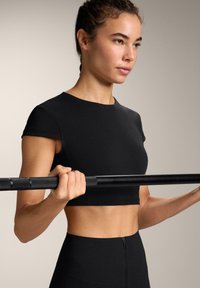 Černý sportovní crop top a legíny s vysokým pasem; model drží černou činku. Hladký materiál s přiléhavým střihem. Minimální design.
