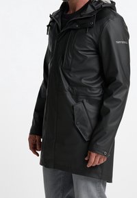 A man models a long black waterproof coat with a hood, featuring large pockets and a front zipper, set against a plain white background.