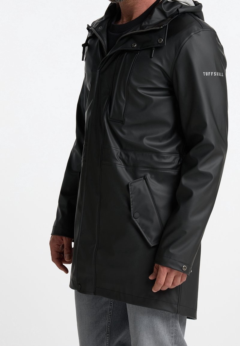 A man models a long black waterproof coat with a hood, featuring large pockets and a front zipper, set against a plain white background.