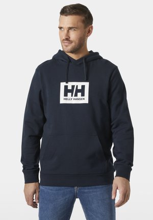 Uomo che indossa una felpa con cappuccio navy Helly Hansen con una tasca frontale e jeans blu, che guarda di lato su uno sfondo semplice.