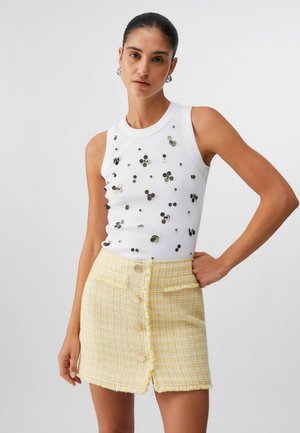 Donna che indossa un top bianco senza maniche con paillettes a fiori e una mini gonna gialla a quadri con bottoni frontali, posando con una mano sul fianco.