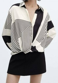 Chemise boutonnée crème et noire avec un motif géométrique en lignes. Associée à une mini-jupe noire. Tissu lisse, coupe décontractée.