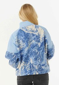Blond persoon met een blauw en wit gevlekte fleecejacket met effen lichtblauwe schouders en zwarte jeans, gezien vanaf de achterkant.