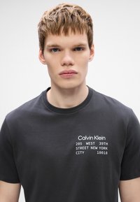 Μαύρη κοντομάνικη βαμβακερή μπλούζα με λευκό κείμενο που λέει "Calvin Klein" και μια διεύθυνση εκτυπωμένη στο μπροστινό μέρος. Στρογγυλή λαιμόκοψη, χαλαρή εφαρμογή.