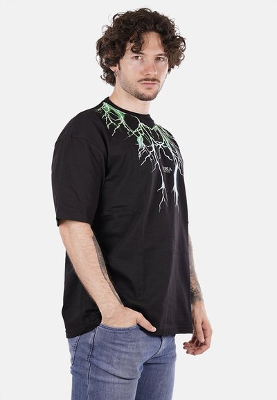 T-shirt in cotone nero con grafiche di fulmini verdi sul collo, maniche corte e una vestibilità rilassata. Indossato con jeans azzurri.