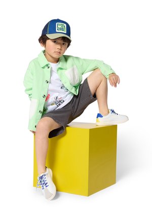 Veste verte boutonnée, t-shirt blanc avec un motif graphique, short gris, baskets bleues et jaunes, posée sur un cube jaune vif.