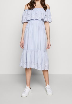Femme en robe midi bleu clair à épaules dénudées avec détails en œillets et ourlet à volants, portant des baskets blanches, debout contre un mur uni.