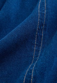 Material de denim albastru cu cusături vizibile în fir de aur; textură netedă cu o ușoară strălucire; plierile stofei creează adâncime și contur.