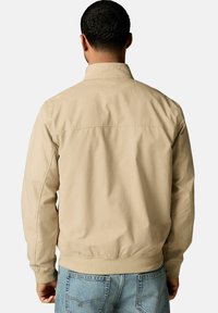 Veste bomber beige avec une texture lisse, poches avant et taille côtelée. Le dos présente une couture horizontale au niveau des épaules.