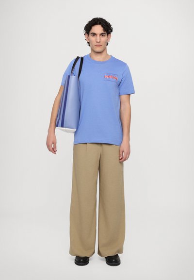 T-shirt en coton bleu clair avec logo "MARNI" rouge, associé à un pantalon large beige et des chaussures noires. Tenant un sac vertical rayé multicolore.