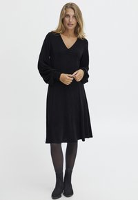 Svart klänning med V-ringning, långa puffärmar och en flödande kjol. Tillverkad av strukturerad textil, kombinerad med svarta ankelstövlar.