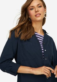 Camisa de botones azul marino de manga larga con un bolsillo, superpuesta sobre una camiseta de rayas moradas y blancas. Textura de tela suave.