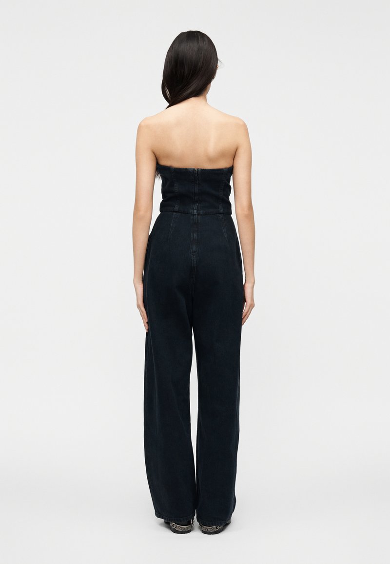 Kvinna stående med ryggen mot kameran, iklädd en svart axelbandslös jumpsuit med vida ben och dragkedja baktill, samt sneakers, på vit bakgrund.