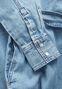 Heledsinine denim-särkide varrukas, millel on kaks hõbedast press-nuppu, topeltõmmatud õmblused ja veidi kulunud tekstuur, rõhutades juhuslikke disainielemente.