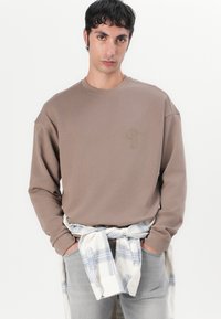 Sudadera marrón de manga larga con puños acanalados y un sutil logo bordado en el pecho. Lleva una camisa de cuadros atada a la cintura.