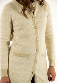 Cardigan largo de crema con un patrón texturizado de una letra. Presenta cuatro botones, dos bolsillos frontales y puños y cuello acanalados.