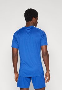 Αθλητική φανέλα σε ζωντανό μπλε ύφασμα, με στρογγυλή λαιμόκοψη, κοντά μανίκια και λευκά στοιχεία στους ώμους. Λείο ύφασμα.