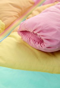 Katoenen beddengoedset met een roze kussen met geplooide randen, rustend op gele en lichtblauwe lakens, die zachte texturen en pastelkleuren toont.