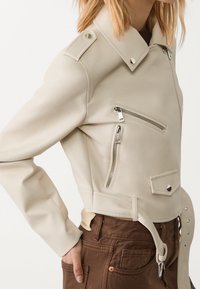 Beżowa kurtka z sztucznej skóry z zamykanymi kieszeniami, naramiennikami, skróconym krojem i detalami w postaci paska. Gładka tekstura i eleganckie akcenty.