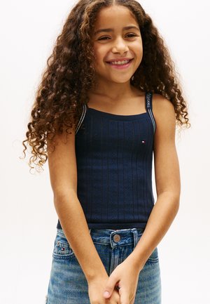 Naeratav tüdruk pikkade lokkis juustega, kes kannab tumesinist kootud topi ja siniseid teksapükse, seisab käed kokku seotud.