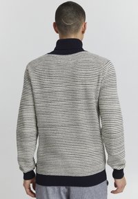 Leichter Strickpullover mit gemusterter Textur, ausgestattet mit hohem Kragen und dunkelblauen Rippbündchen sowie -saum. Farbgebung in Creme und Marineblau.