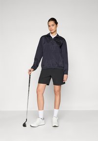 Marineblauwe golfpull met lange mouwen en een halfrits, elastische zoom, gecombineerd met zwarte shorts en witte sneakers. Het model houdt een golfclub vast.