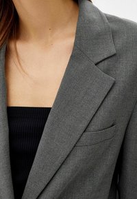 Frau mit einem grauen Blazer über einem schwarzen, gerippten Oberteil, das den Ausschnitt sowie einen Teil des Revers und die Brusttasche des Blazers zeigt.