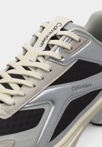 Αθλητικό παπούτσι Calvin Klein με μπεζ κορδόνια, γκρι και μαύρες επιφάνειες, και μεταλλικές ασημί λεπτομέρειες, εμφανίζοντας πλαϊνές και πάνω όψεις.