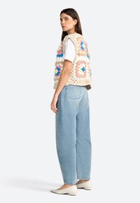 Crocheted vest with pastel squares in blue, pink, and cream, paired with light blue jeans and white pointed flats. Model posed from side.