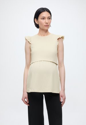 Tehotná žena s krátkymi tmavými vlasmi, oblečená v béžovom tehotenskom topu s volánmi na rukávoch a čiernych nohaviciach, stojaca pred jednofarebným pozadím.