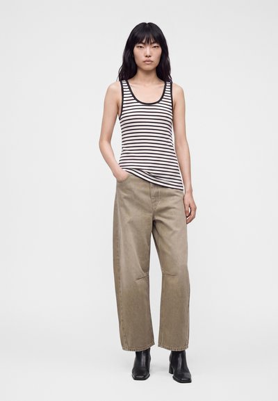 Femme aux longs cheveux noirs portant un débardeur rayé noir et blanc, un pantalon large beige et des bottines noires, debout avec une main dans la poche.