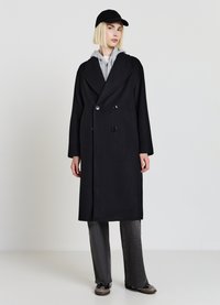 Cappotto doppiopetto nero in misto lana, caratterizzato da grandi bottoni e colletto a risvolto. Abbinato a una felpa con cappuccio grigia, pantaloni grigi e un berretto nero.