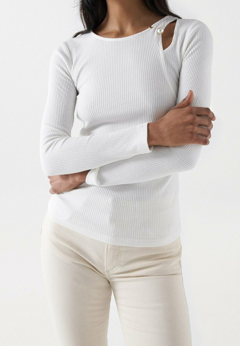 Femme portant un haut blanc à manches longues côtelé avec un détail de boutons près de l'épaule gauche et un pantalon beige clair, bras croisés.