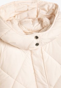 Gepolsterte, hellbeige Pufferjacke mit hohem Kragen. Verfügt über einen Druckknopfverschluss und ein glattes, glänzendes Material mit weicher Textur.