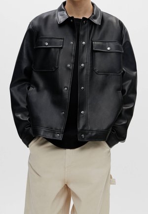 Personne portant une veste noire en similicuir boutonnée avec des poches poitrine à rabat et un pantalon beige, debout les mains dans les poches.