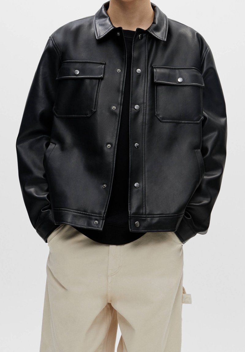 Personne portant une veste noire en similicuir boutonnée avec des poches poitrine à rabat et un pantalon beige, debout les mains dans les poches.