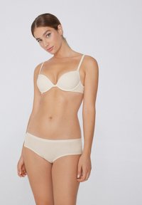 Beige bezšvová podprsenka s kosticemi a nastavovacími ramínky, v kombinaci s ladícími béžovými šortkami. Oba kusy mají jemnou a hladkou texturu.