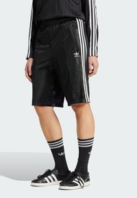 Zwarte sportshorts met drie witte strepen aan de zijkanten, met een subtiele glans en een Adidas-logo op de linker zoom. Model gecombineerd met gestreepte sokken.