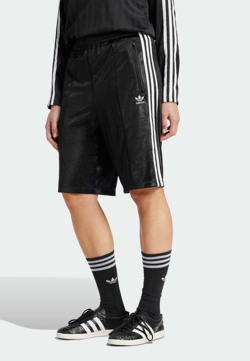 Zwarte sportshorts met drie witte strepen aan de zijkanten, met een subtiele glans en een Adidas-logo op de linker zoom. Model gecombineerd met gestreepte sokken.