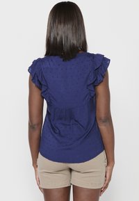 Marineblaue Bluse aus leichtem Stoff, mit voluminösen kurzen Ärmeln, strukturierten Punkten und einer lockeren Passform im Rücken. Kombiniert mit khakifarbenen Shorts.