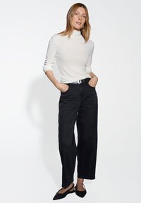 Femme portant un col roulé blanc, un jean large noir avec une ceinture noire, et des ballerines à talon noir, se tenant les mains dans les poches.