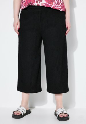 Czarne, plisowane spodnie szerokie z teksturowanej tkaniny, z talią na wysokości średniej i luźnym krojem, w połączeniu z białymi klapkami bez zapinania.