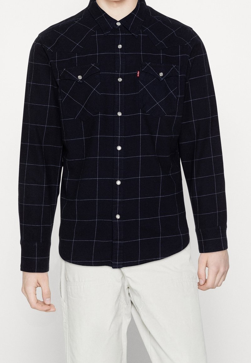 Chemise en flanelle bleu marine à manches longues avec motif à carreaux blanc, col boutonné, deux poches poitrine et boutons-pression argentés.
