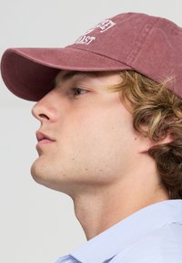 Cappellino da baseball bordeaux con testo ricamato, materiale in cotone, visiera curvata, corona piatta, indossato con una camicia azzurro chiaro a collo.