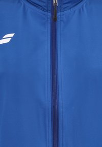 Blå jakke med central lynlås og subtilt logo på venstre side nær kraven, der viser stoffets tekstur og syningsdetaljer.