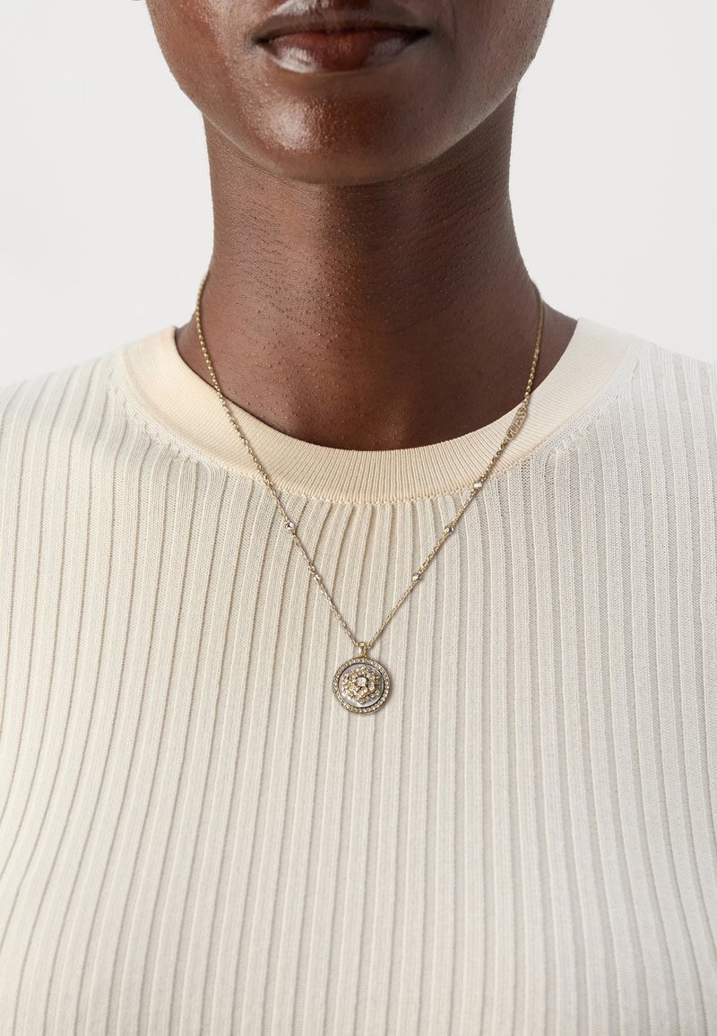 Gros plan d'une personne portant un pull crème côtelé et un collier chaîne en or avec un pendentif rond serti de diamants.