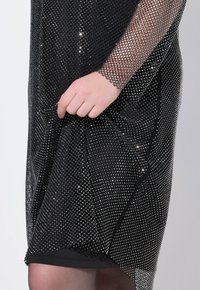 Schwarzes Netzkleidungsstück mit funkelnden silbernen Akzenten, das ein lockeres Design und eine netzartige Textur aufweist und über einer weichen schwarzen Schicht darunter hängt.